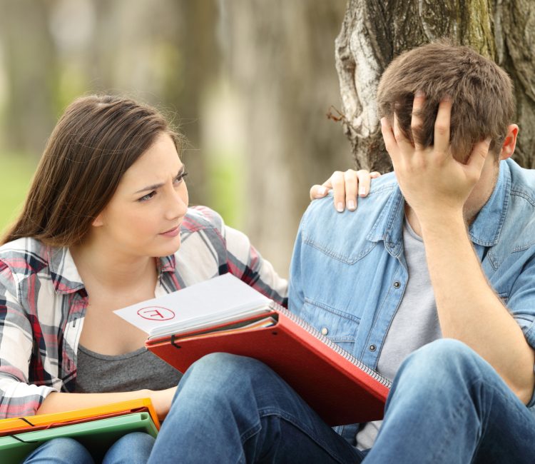 Friend comforting to a sad student with failed exam sitting on the grass in a park