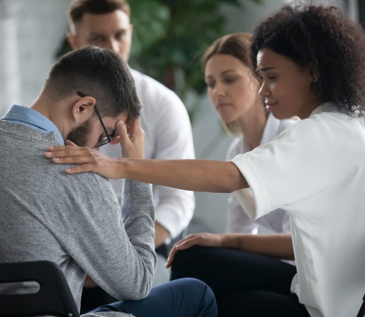 Caring African American woman support comfort unhappy sad Caucasian male friend at counseling meeting indoors. Supportive ethnic female cheer caress upset depressed man suffering from problem.